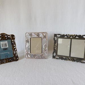 May include: Three metal picture frames: a bronze-toned frame with cherub motifs, a silver-toned frame with rose accents, and a rectangular bronze-toned double frame with ornate scrollwork edges. The bronze frame includes a 3x5 inch label.