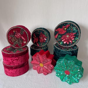 Op de afbeelding: Een set van zes kerstversierde decoratieve dozen. De dozen zijn gemaakt van fluweel en hebben verschillende kerstontwerpen, waaronder kardinalen, kerststerren en hulst. De dozen zijn in tinten rood en groen.