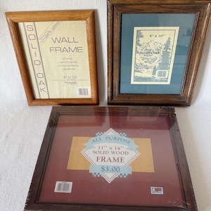 May include: Three wooden picture frames. The top left frame is light brown and reads "SOLID OAK WALL FRAME 8 x 10". The top right frame is dark brown with a blue mat and a label that reads "8" x 10" American Oak Frame". The bottom frame is dark brown and reads "ALL PURPOSE 11" x 14" SOLID WOOD FRAME".