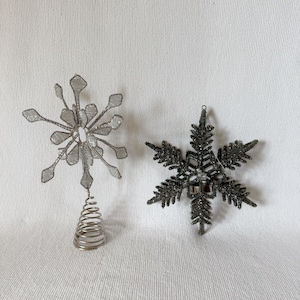 Puede incluir: Dos adornos decorativos de copos de nieve. Uno es plateado con un diseño de alambre y una base de resorte. El otro es un copo de nieve gris oscuro y brillante. Ambos están diseñados para la decoración navideña.