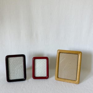 Puede incluir: Tres marcos de fotos rectangulares dispuestos en fila. El marco de la izquierda es de color marrón oscuro, el marco central es de un rojo vibrante y el marco de la derecha es de un tono madera claro. Cada marco tiene un diseño de bordes lisos.
