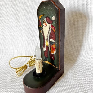 May include: A wooden Santa Claus candle holder with a green and brown painted finish. The Santa Claus is holding a bag of gifts and a candle. The candle holder has a light bulb that illuminates the candle.