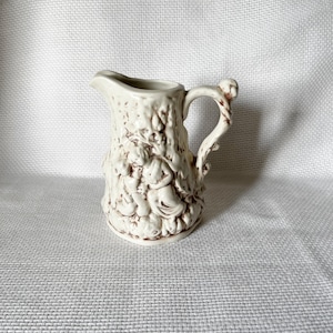 May include: A white ceramic creamer with a detailed design of a couple in a tree. The creamer has a handle shaped like a tree branch.