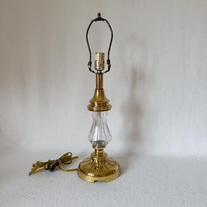 Puede incluir: Una lámpara de mesa vintage con base y detalles de latón. La lámpara presenta una sección central de vidrio transparente y acanalado y un soporte de pantalla negro. La lámpara tiene un cable de alimentación dorado y está sobre un fondo blanco.