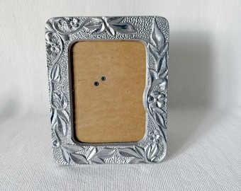 May include: A silver picture frame with a floral design. The frame is made of metal and has a textured surface.