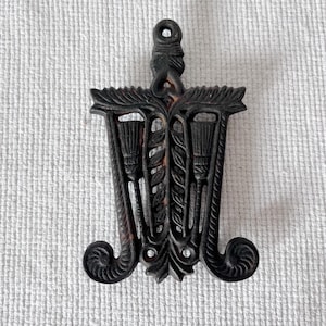 May include: Black cast iron trivet with a decorative design of three stylised wheat stalks. The trivet has a loop at the top for hanging.