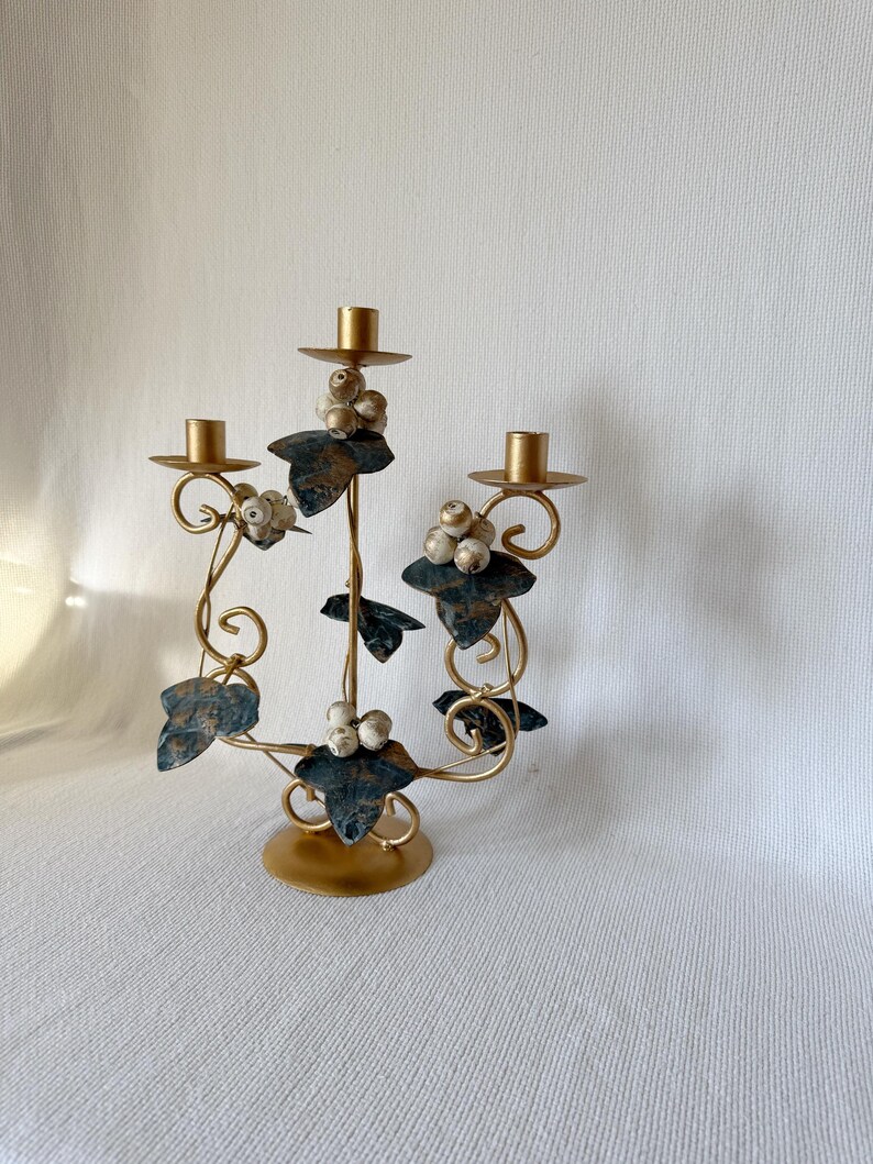 Puede incluir: Un candelabro de metal dorado con tres candeleros. El candelabro presenta hojas decorativas con detalles en azul-verde y racimos de bayas blancas. La base es redonda y el dise&ntilde;o general es ornamentado e inspirado en lo vintage.