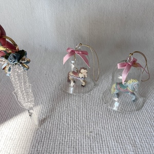 May include: Three glass bell ornaments with pink ribbons and a carousel horse figurine inside each. The horses are painted in pastel colors.