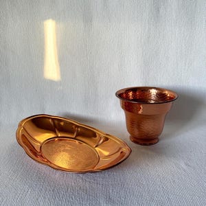 May include: A copper-coloured, hammered metal planter and a decorative dish. The planter has a flared rim and a textured surface. The dish is oval-shaped with a raised rim and a detailed centre design. Both items have a shiny finish.