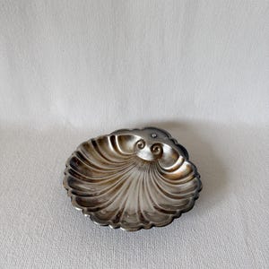 May include: A silver-toned, shell-shaped dish with a detailed, ridged design. The dish has a scalloped edge and a slightly tarnished appearance, suggesting age. The dish is likely for decorative use.
