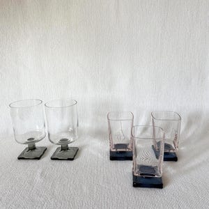 May include: A collection of six clear glass drinking glasses. Two glasses have a rounded shape and dark gray square bases. The other four glasses have a square shape with a pink hue and dark blue square bases.