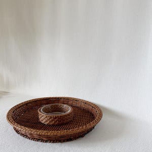 May include: A round, woven brown rattan serving tray with a smaller, matching bowl in the center. The tray has a raised edge and a textured surface, perfect for serving snacks or appetizers. The background is a plain white.