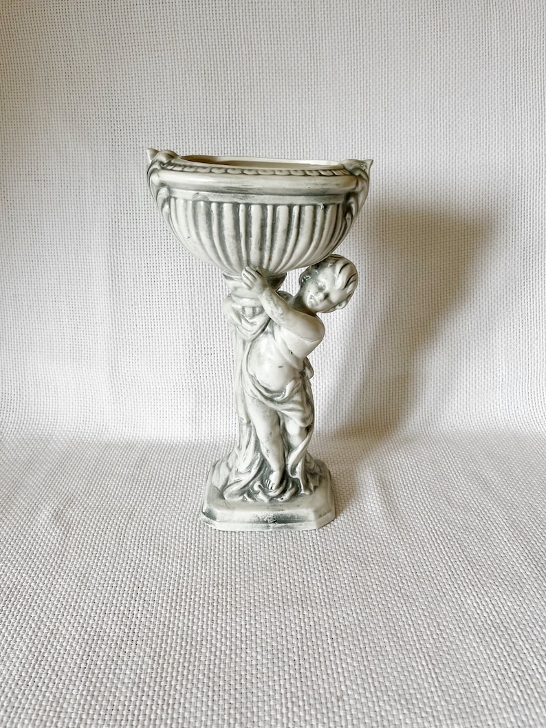 May include: A white ceramic planter with a cherub holding up a round bowl. The planter has a distressed finish and a square base.