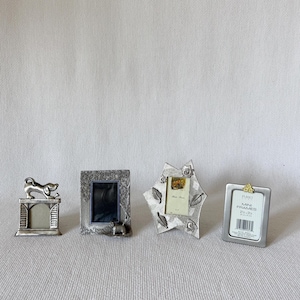 May include: Four small metal picture frames. One features a dog on a fireplace mantle, one has a textured tree design with a dog, one is star-shaped with leaf accents, and one is a simple rectangular frame with the text MINI FRAMES 2.5 x 3.5.