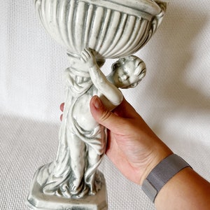 May include: A white ceramic planter shaped like a cherub holding a bowl. The planter has a gray glaze and is in the style of a classical sculpture.