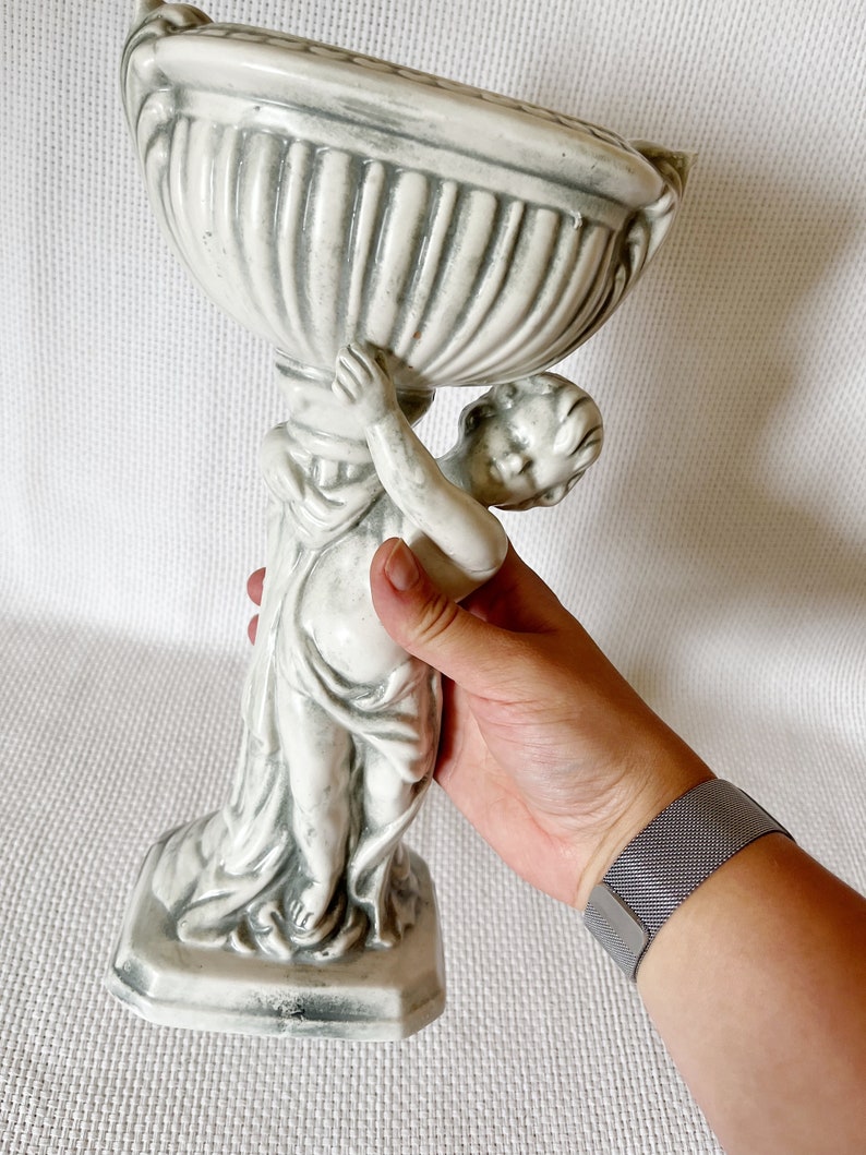 May include: A white ceramic planter shaped like a cherub holding a bowl. The planter has a gray glaze and is in the style of a classical sculpture.