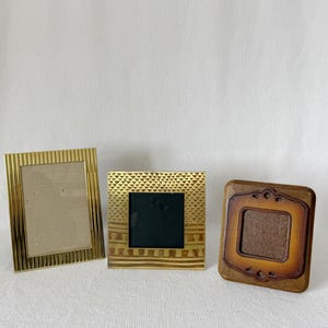 May include: Three decorative picture frames. The frames are in gold and brown tones. The gold frames have textured designs, while the brown frame has a carved, ornate design. The frames are empty, ready for photos.