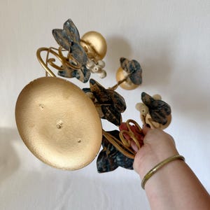 Puede incluir: Un candelabro de metal dorado con base redonda y detalles decorativos florales y de hojas. Las hojas son una mezcla de oro, azul y marr&oacute;n. El candelabro tiene un dise&ntilde;o caprichoso y art&iacute;stico.