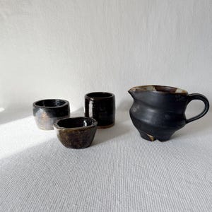 May include: A collection of handmade ceramic tableware. The set includes a small creamer with a handle, two small cylindrical cups, and a small bowl. The pieces are glazed in shades of black and brown, with a textured finish.