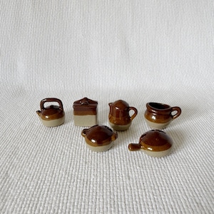 May include: A set of six miniature ceramic kitchen items featuring a two-tone brown and beige glaze. The collection includes a small teapot, a square container, a pitcher, a mug, and two small lidded pots with handles, all arranged together.