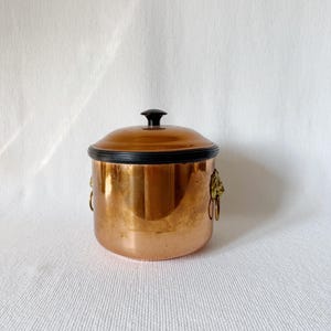 Puede incluir: Una olla de color cobre con tapa. La olla tiene forma redonda con borde negro y pomo negro en la tapa. Cuenta con asas decorativas con cabeza de león y anillas. La olla está sobre un fondo blanco.