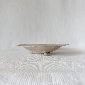 Puede incluir: Un cuenco poco profundo de metal plateado de forma circular, con un borde decorativo con motivos florales en relieve. El cuenco se apoya sobre tres pequeños pies esféricos, proporcionando una base estable y un aspecto bajo y elegante.