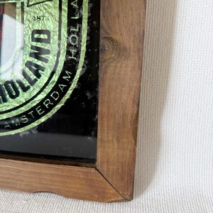 Puede incluir: Letrero enmarcado en madera con el logotipo de Heineken. El letrero presenta un dise&ntilde;o de l&aacute;mina verde y dorada con las palabras "Amsterdam Holland" y el a&ntilde;o "1873". El marco es de madera marr&oacute;n claro.