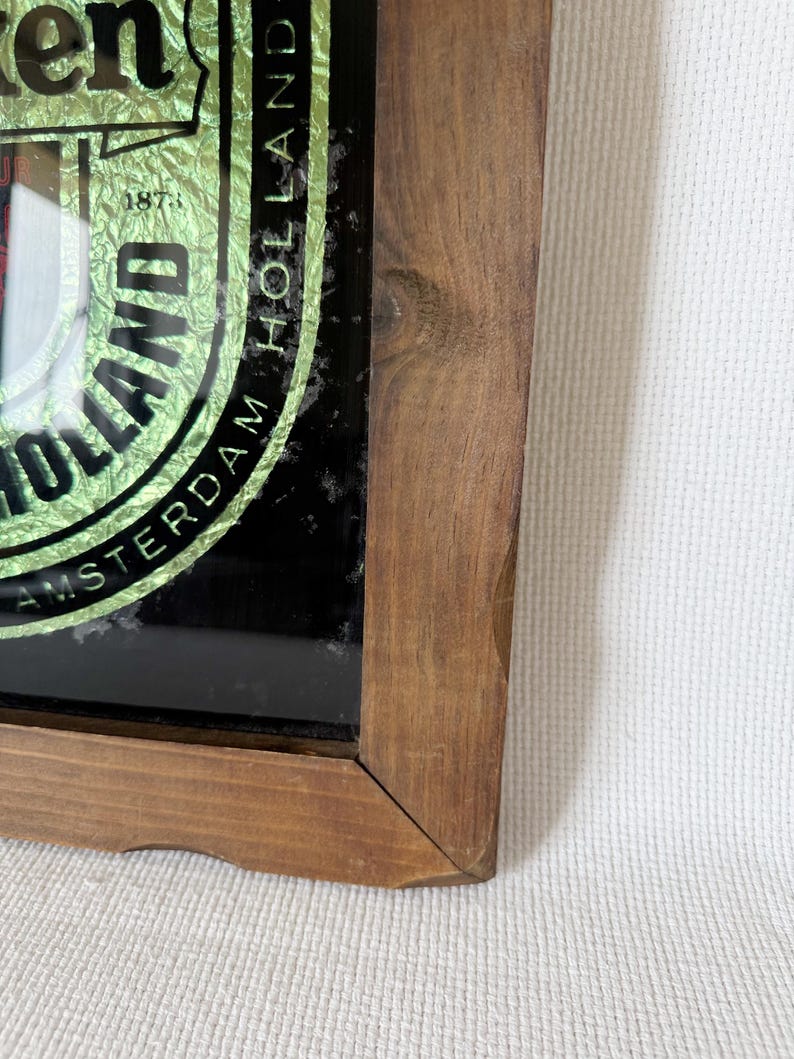 Puede incluir: Letrero enmarcado en madera con el logotipo de Heineken. El letrero presenta un dise&ntilde;o de l&aacute;mina verde y dorada con las palabras "Amsterdam Holland" y el a&ntilde;o "1873". El marco es de madera marr&oacute;n claro.