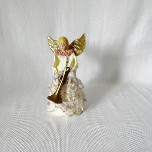 May include: A decorative angel figurine holding a trumpet. The angel has blonde hair, gold wings, and a cream-coloured dress with lace and rose accents. The trumpet is gold-coloured.