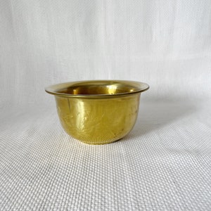 Puede incluir: Un pequeño cuenco de metal dorado con un borde ligeramente acampanado y un diseño floral grabado. El cuenco está sobre una superficie blanca texturizada.