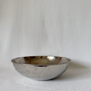 May include: A silver-colored bowl with a hammered texture. The bowl has a rounded shape and a slightly flared rim. The interior of the bowl has a reflective surface. The bowl is set against a white background.