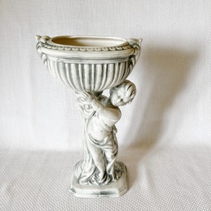 May include: A white ceramic planter with a cherub holding up a bowl. The planter has a gray and white striped design.