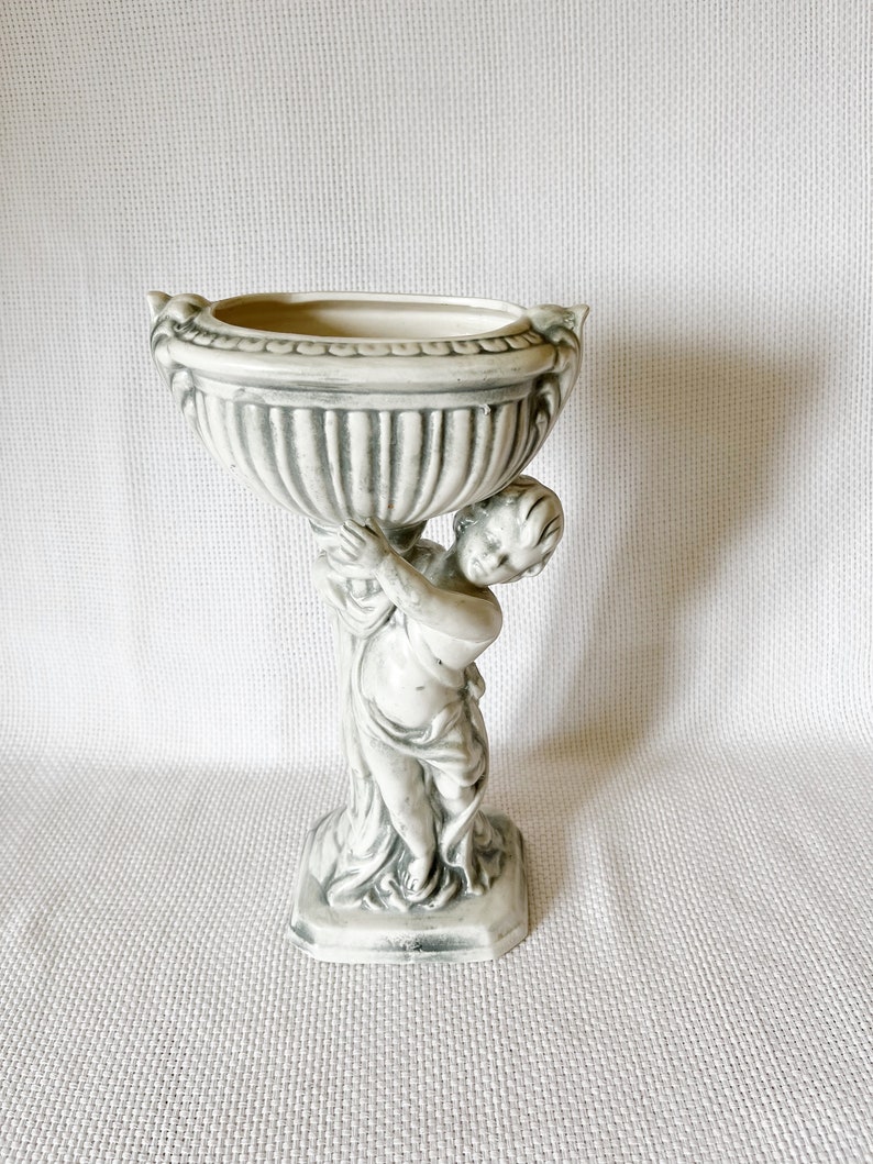 May include: A white ceramic planter with a cherub holding up a bowl. The planter has a gray and white striped design.