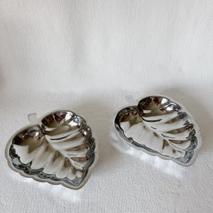 May include: Two silver-colored leaf-shaped dishes. The dishes have a reflective, metallic finish and are placed on a white textured surface. The leaf design features detailed veining, adding to their realistic appearance. These dishes could be used for serving small portions.