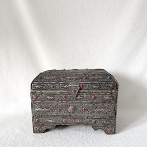May include: Ornate, antique silver chest with a curved top. The chest is decorated with red and blue gemstones and intricate metalwork. The chest has a small latch and is likely used for storing jewelry or small items.
