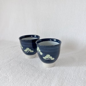 May include: Two blue ceramic teacups with white and green floral designs. The cups have a white rim and base.