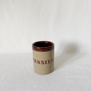 May include: A brown and beige ceramic utensil holder with the word "UTENSILS" printed in brown on the front.