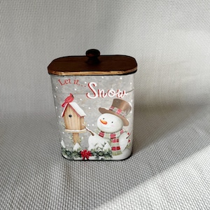 May include: A metal tin container with a wooden lid. The tin features a snowman with a red and black scarf, a red cardinal, and a birdhouse. The text "Let it... Snow" is printed on the tin.