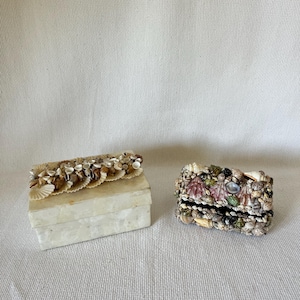 May include: Two decorative boxes made of white material with a seashell design. One box is rectangular and the other is square. Both boxes are decorated with a variety of seashells.