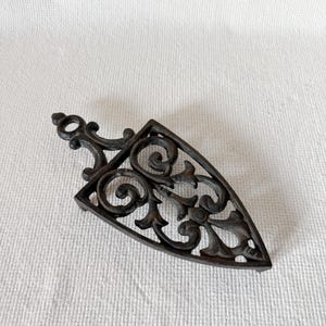 May include: A black cast iron trivet with an ornate, openwork design. The trivet is shaped like an iron, with a handle and a triangular base. The decorative pattern includes swirls and floral elements, adding a vintage aesthetic.
