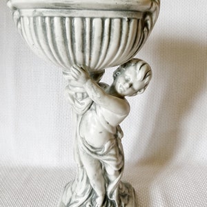 May include: A white ceramic planter with a cherub holding up the planter. The planter has a fluted design and a square base.