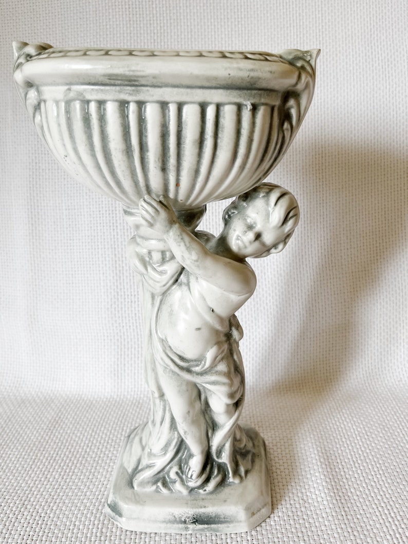 May include: A white ceramic planter with a cherub holding up the planter. The planter has a fluted design and a square base.