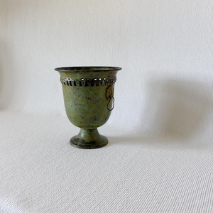May include: A weathered green metal urn with a pedestal base and a flared rim featuring a decorative cutout pattern. The vessel has a distressed finish and includes a small metal ring handle attached to the side, showcasing a textured appearance.
