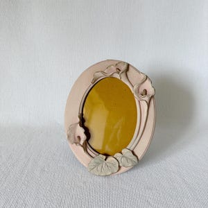 Könnte beinhalten: Ovaler, hellrosa Bilderrahmen mit floralem Design. Der Rahmen hat Akzente von Calla-Lilien und Blättern. Das Innere des Rahmens ist senfgelb.
