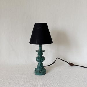 Könnte beinhalten: Eine kleine türkisfarbene Tischlampe mit schwarzem Lampenschirm. Die Lampe hat einen dekorativen Sockel mit Zierdetails und ein schwarzes Netzkabel. Die Lampe ist etwa 30 cm hoch.