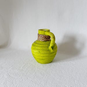 Puede incluir: Un pequeño jarrón de cerámica verde brillante con una superficie texturizada y un asa de tejido marrón. El jarrón está sobre un fondo blanco.