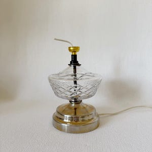 Puede incluir: Una lámpara de mesa de vidrio transparente con una base dorada y plateada. La lámpara tiene un patrón de diamante en la pantalla de vidrio.