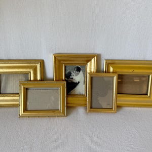 以下が含まれることがあります： さまざまなサイズの金色の写真立てが5つ集まったもの。1つのフレームには白黒写真が入っています。フレームは白い表面に配置され、装飾的で長方形のデザインが特徴です。大切な思い出を飾るのに最適です。