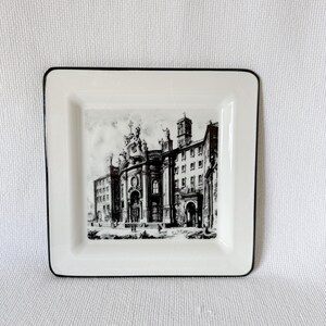 May include: A square ceramic plate with a white base and a thin black rim. The center features a detailed black and white illustration of a historic European cathedral with ornate architectural details, arched doorways, and a tall bell tower.
