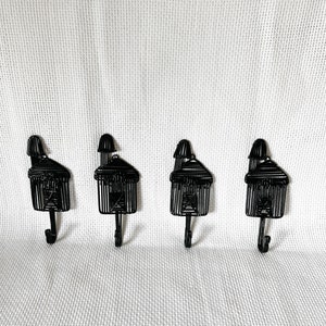 May include: Four black metal hooks shaped like houses with a small roof on top. The hooks are mounted on a white wall.